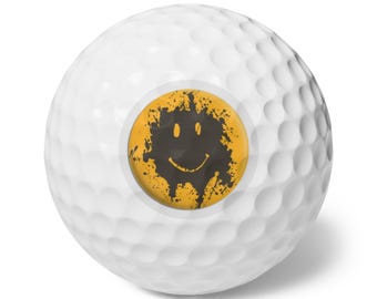 May include: White golf ball featuring a yellow and black smiley face design. The smiley face has a distressed, splatter paint effect. The golf ball has a dimpled surface.
