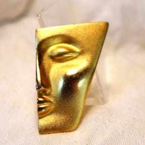May include: A gold-toned metal brooch in the shape of a stylized face. The face is in profile, with one eye closed and the mouth slightly open.