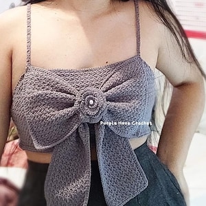 Op de afbeelding: Een grijs gehaakt topje met een grote strik en een bloemdetail. Het topje is kort en heeft dunne bandjes. De tekst "Purple Haze Crochet" is zichtbaar op de voorkant van het topje.