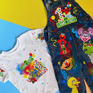 May include: A white t-shirt and blue denim overalls featuring colorful Sesame Street character graphics. The designs include Elmo, Big Bird, Bert, and Abby Cadabby, with the text "LIVANS STREET" and a large number one, accented by vibrant paint splatters.