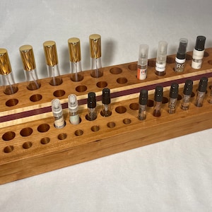 May include: A wooden organizer with multiple holes to hold small perfume bottles. The organizer has a light brown wood finish with a purple and white inlay. There are 10 gold-capped perfume bottles and 15 black-capped perfume bottles in the organizer.