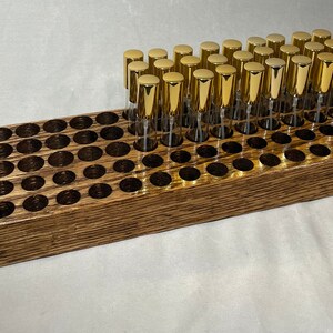 May include: A wooden storage rack with 48 round holes for holding small bottles. The rack is made of dark stained wood and has a natural wood grain. There are 30 gold-capped glass bottles in the rack.