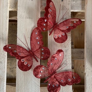 May include: Two red glitter butterflies with silver accents on a wooden background.