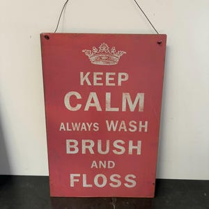 Könnte beinhalten: Rechteckiges rotes Holzschild mit weißem Text "KEEP CALM ALWAYS WASH BRUSH AND FLOSS". Oben befindet sich eine weiße Kronengrafik. Das Schild wird von einem schwarzen Draht gehalten.