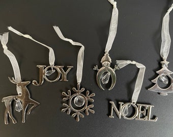 Könnte beinhalten: Sechs silberne Weihnachtsbaumschmuckstücke mit weißen Schleifen. Die Schmuckstücke zeigen ein Rentier, eine Schneeflocke, das Wort "JOY", einen Schneemann, einen Weihnachtsbaum und das Wort "NOEL". Jedes Schmuckstück hat einen klaren Kristall in der Mitte.