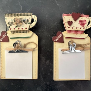 Puede incluir: Dos portapapeles decorativos con diseños de tazas de té. Una taza de té tiene un patrón a cuadros verde y blanco, la otra tiene un patrón a cuadros rojo y blanco. Cada portapapeles tiene una etiqueta de "Bienvenida" en forma de corazón.