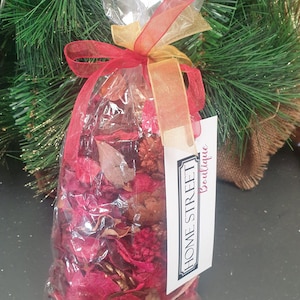 Op de afbeelding: Een doorzichtige plastic zak gevuld met rode, bruine en gouden potpourri. De zak is vastgebonden met een rood en gouden lint en heeft een label met de tekst "Home Street Boutique".