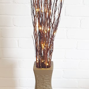 May include: A decorative floor vase with a twisted design, filled with brown branches and small, warm white LED lights. The vase is a neutral tan color and sits on a wooden pallet.