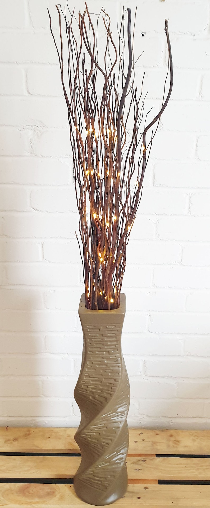 May include: A decorative floor vase with a twisted design, filled with brown branches and small, warm white LED lights. The vase is a neutral tan color and sits on a wooden pallet.