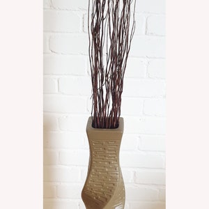 May include: A tall, taupe-colored vase with a textured, twisted design. The vase is filled with a bundle of dark brown, slender branches. The background is a white brick wall.