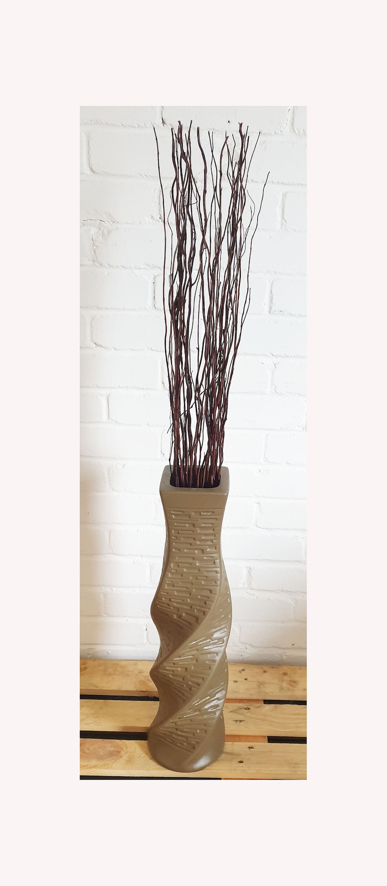 May include: A tall, taupe-colored vase with a textured, twisted design. The vase is filled with a bundle of dark brown, slender branches. The background is a white brick wall.