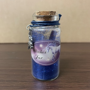Puede incluir: Un pequeño frasco de vidrio lleno de purpurina azul con una tapa de corcho. El frasco tiene una etiqueta que dice "Wishes Jar" y presenta un unicornio blanco con estrellas.