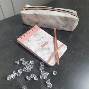Op de afbeelding: Een set met een etui en notitieblok met marmerpatroon en roségouden accenten. Het notitieblok heeft een roze bovenkant en de tekst 'NOTES'. Een roségouden pen en decoratieve kristallen maken de set compleet.