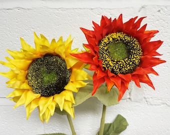 Pode incluir: Dois girassóis artificiais, um amarelo e outro vermelho, com centros verdes e folhas. Os girassóis estão sobre um fundo de parede de tijolo branco.