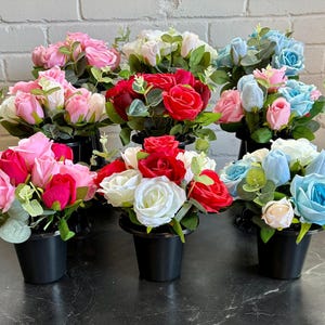 Può includere: Disposizione di rose artificiali in vasi neri. Le rose sono in vari colori, tra cui rosa, rosso, bianco e blu, con fogliame verde. I vasi sono disposti su una superficie scura contro un muro di mattoni bianchi.