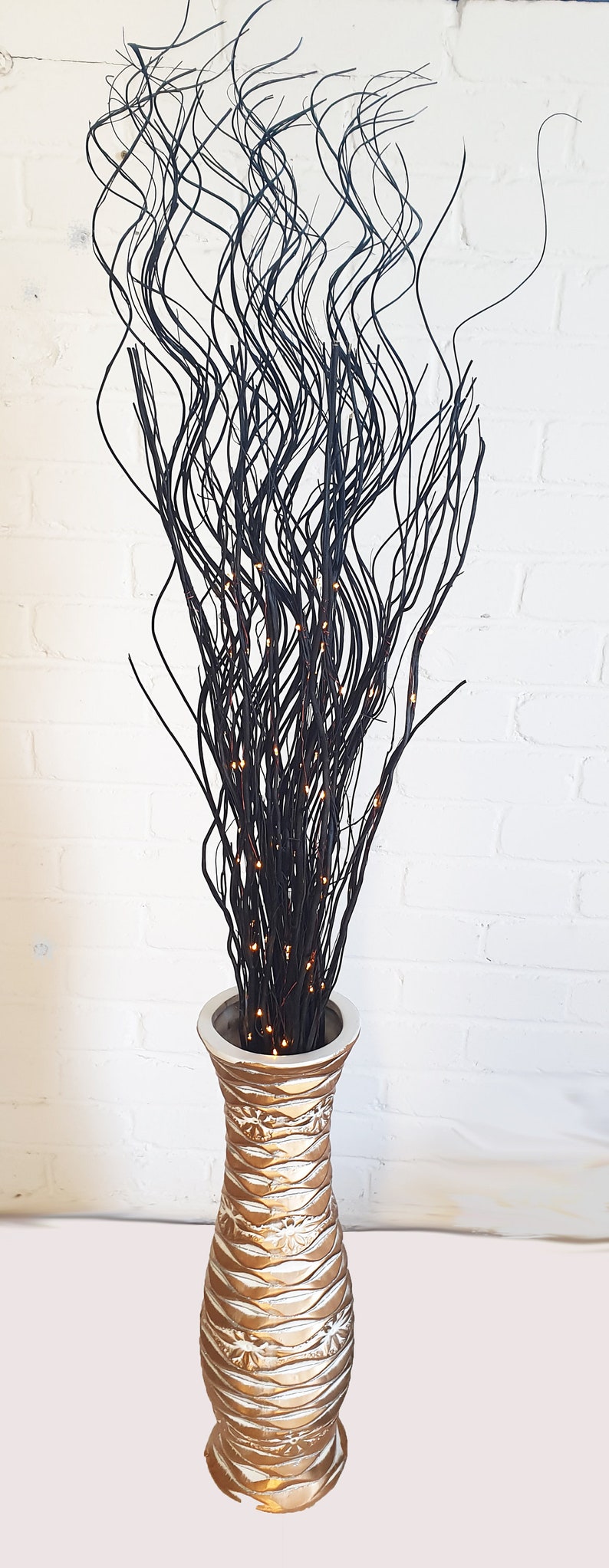 May include: A tall, gold vase with a textured surface holds a bouquet of black curly willow branches with small, warm white lights.