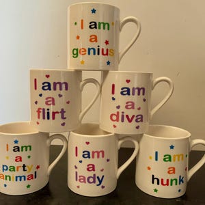 Peut inclure: Tasses en céramique blanches avec des phrases colorées telles que "I am a genius", "I am a flirt" et "I am a diva". Chaque tasse a une anse incurvée et est décorée d'étoiles et de coeurs.