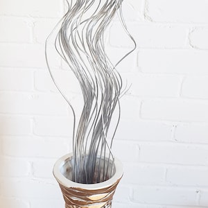May include: A tall, gold and white ceramic vase with a wavy, textured design. The vase is filled with long, silver, curly stems.