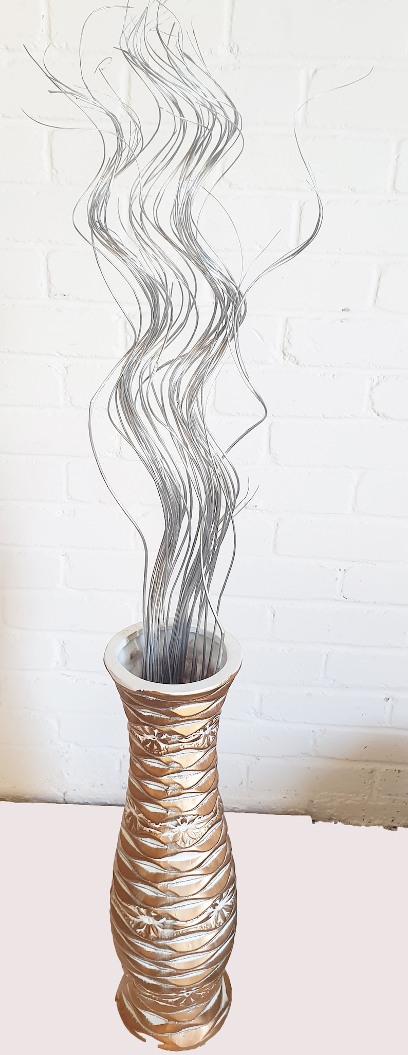May include: A tall, gold and white ceramic vase with a wavy, textured design. The vase is filled with long, silver, curly stems.