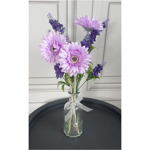 May include: A bouquet of lavender and light purple gerbera daisies in a clear glass vase with a white ribbon.