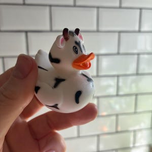 Peut inclure: Un canard en caoutchouc blanc avec des taches noires, des cornes marron et un bec orange. Le canard est con&ccedil;u pour ressembler &agrave; une vache. Le canard est tenu dans une main sur un fond carrel&eacute; blanc.