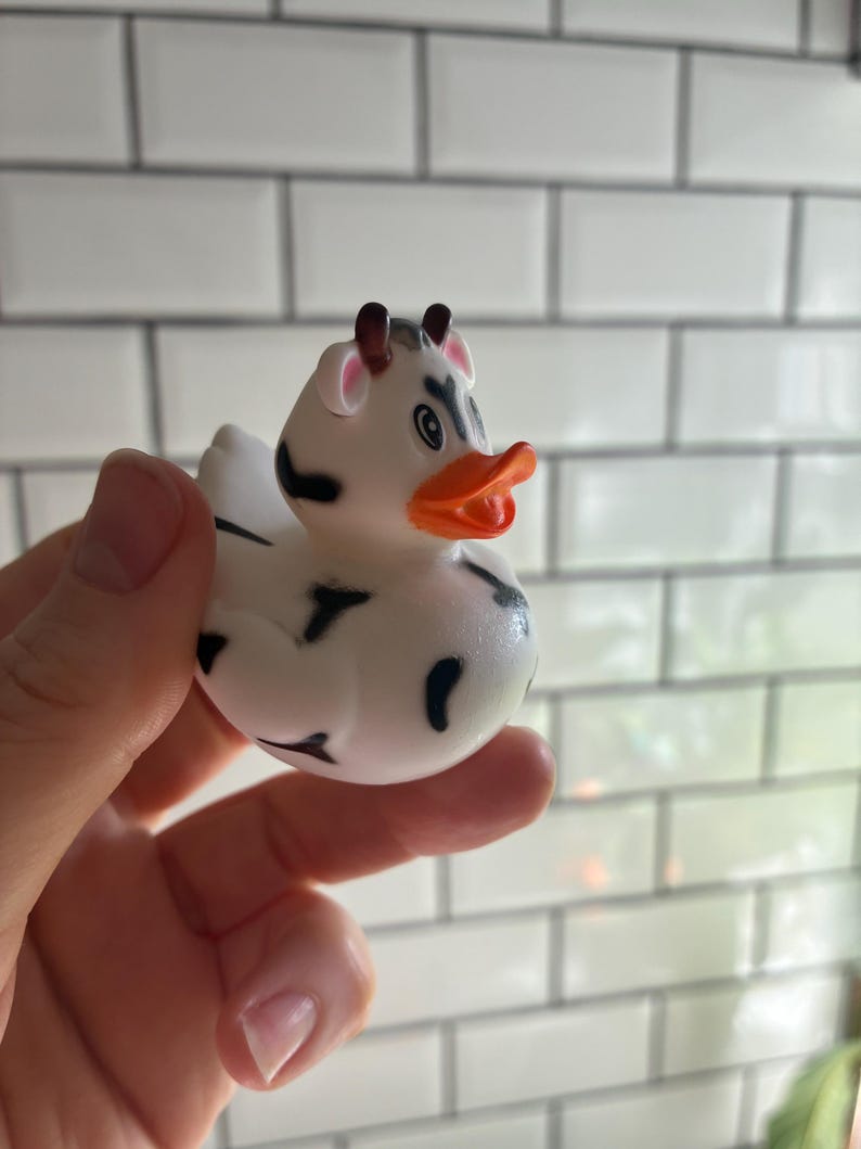 Peut inclure: Un canard en caoutchouc blanc avec des taches noires, des cornes marron et un bec orange. Le canard est con&ccedil;u pour ressembler &agrave; une vache. Le canard est tenu dans une main sur un fond carrel&eacute; blanc.