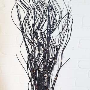 May include: Black twig branches with warm white LED lights in a gold and white vase. The branches are arranged in a wavy pattern.