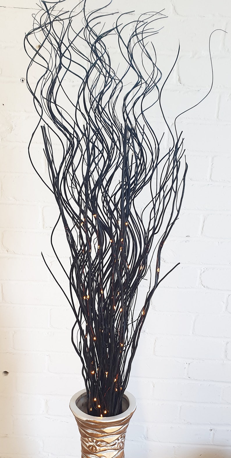 May include: Black twig branches with warm white LED lights in a gold and white vase. The branches are arranged in a wavy pattern.