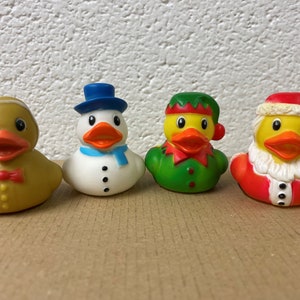 Peut inclure: Quatre canards en caoutchouc v&ecirc;tus de costumes de f&ecirc;te. L'un est un bonhomme en pain d'&eacute;pices, un bonhomme de neige avec un chapeau et une &eacute;charpe bleus, un elfe en vert et un P&egrave;re No&euml;l avec un costume et un chapeau rouges.