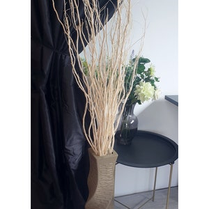 Puede incluir: Un jarrón alto de cerámica marrón retorcido lleno de ramas de ramitas blancas. El jarrón está sobre un suelo de madera delante de una cortina negra.