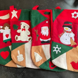 May include: Four red and green Christmas stockings with different embroidered designs. The stockings feature a snowman, a teddy bear, an angel, and Santa Claus.