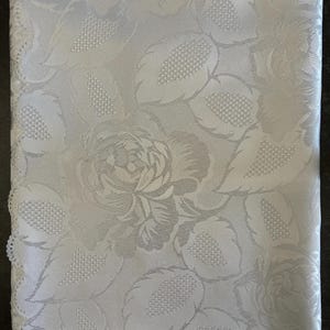 Könnte beinhalten: Eine weiße Tischdecke aus Stoff mit einem Ton-in-Ton Jacquard-Gewebe, das ein sich wiederholendes Muster aus großen blühenden Rosen und detaillierten Blättern zeigt. Das Textil hat einen subtilen Glanz und eine strukturierte Oberfläche mit Spitzenrand.