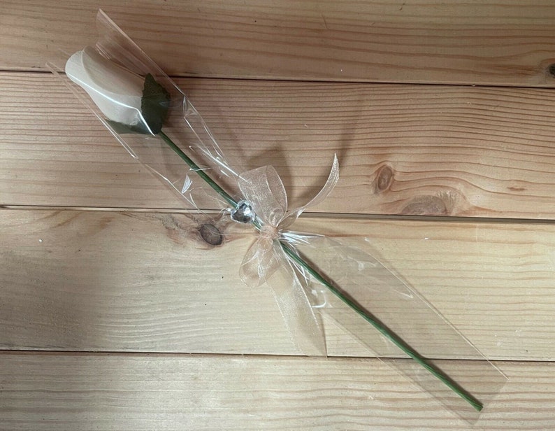 May include: A single white rose wrapped in clear plastic with a beige ribbon and a silver heart charm.