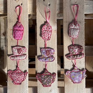 Puede incluir: Tres adornos de tela rosa y blanca en forma de taza de té y tetera colgando de una superficie de madera. Cada adorno presenta un diseño diferente, incluyendo una tetera, una taza de té y una magdalena.