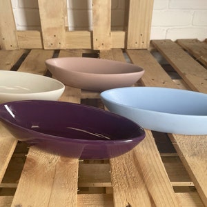 Op de afbeelding: Vier keramische serveerschalen in verschillende kleuren: wit, lichtroze, lichtblauw en paars. De schalen zijn ovaalvormig en hebben een glanzende afwerking.