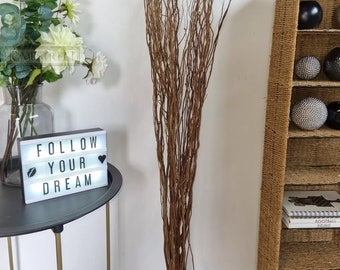 May include: A bunch of brown willow branches, tied together at the bottom, against a white wall. The branches are long and thin, and they are arranged in a loose, natural way. The branches are a popular choice for floral arrangements and home decor.