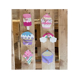 May include: Six wooden wall hooks shaped like ice cream cones, cupcakes, and a rainbow. The hooks are painted in pastel colors with pink, purple, and yellow accents.