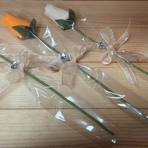 May include: Three wooden roses wrapped in clear plastic with a beige ribbon and a silver charm. The roses are yellow, orange, and white.