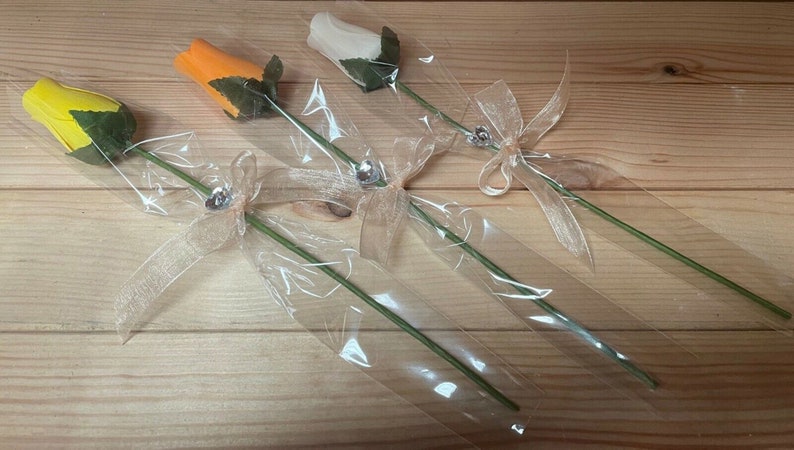 May include: Three wooden roses wrapped in clear plastic with a beige ribbon and a silver charm. The roses are yellow, orange, and white.