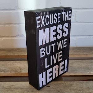 Puede incluir: Un letrero de madera negro con texto blanco que dice "Disculpe el desorden, pero vivimos aquí!"