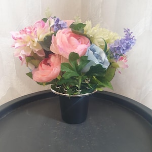 Pode incluir: Um arranjo floral com flores artificiais rosa, azuis e lavanda num vaso preto com uma borda prateada. As flores estão dispostas numa bandeja cinza escuro.