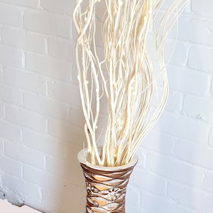 May include: A tall, gold and white ceramic vase with a textured surface, filled with white twig branches.