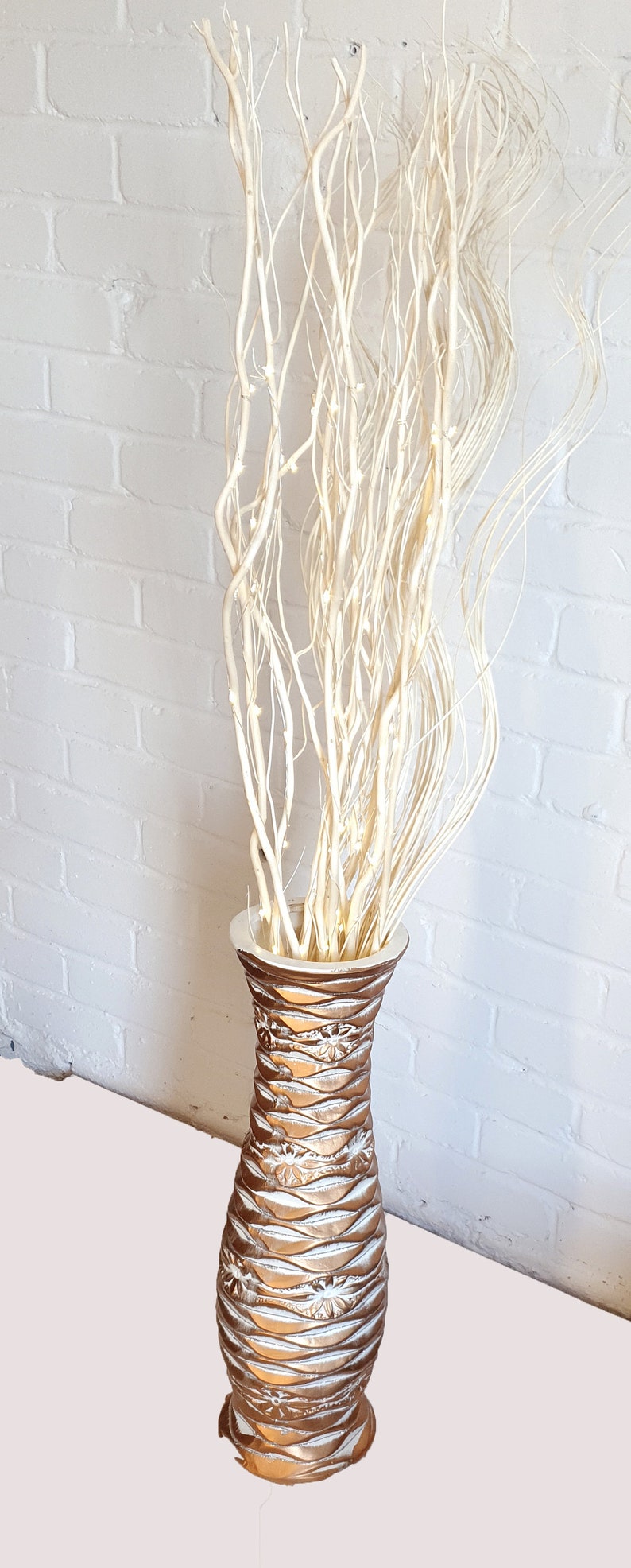 May include: A tall, gold and white ceramic vase with a textured surface, filled with white twig branches.