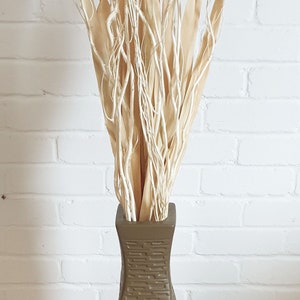 May include: A decorative vase with a twisted design, filled with dried beige and white branches. The vase is a muted olive green color and sits against a white brick wall.