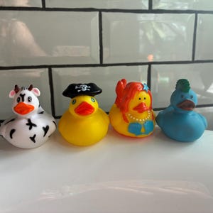 Peut inclure: Quatre canards en caoutchouc color&eacute;s. Un canard vache blanc et noir, un canard pirate jaune, un canard sir&egrave;ne jaune et un canard bleu avec une cr&ecirc;te verte. Tous ont un bec orange.