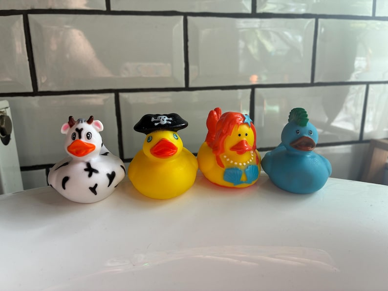 Peut inclure: Quatre canards en caoutchouc color&eacute;s. Un canard vache blanc et noir, un canard pirate jaune, un canard sir&egrave;ne jaune et un canard bleu avec une cr&ecirc;te verte. Tous ont un bec orange.
