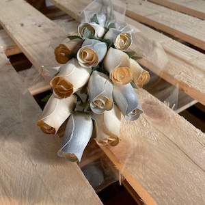 May include: A bouquet of wooden roses in shades of beige and silver, tied with a sheer ribbon. The roses are arranged in a fan shape and are displayed on a wooden surface.