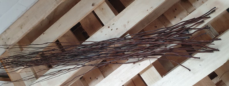 May include: A bundle of thin, dark brown twigs, possibly willow or similar, arranged on a light-colored wooden pallet. The twigs are long and slender, with a natural, organic appearance. The wood grain of the pallet is visible.
