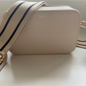 May include: A beige crossbody bag with a black and white striped strap. The bag has a gold zipper and a gold metal clasp. The initials "NH" are embossed on the front of the bag.