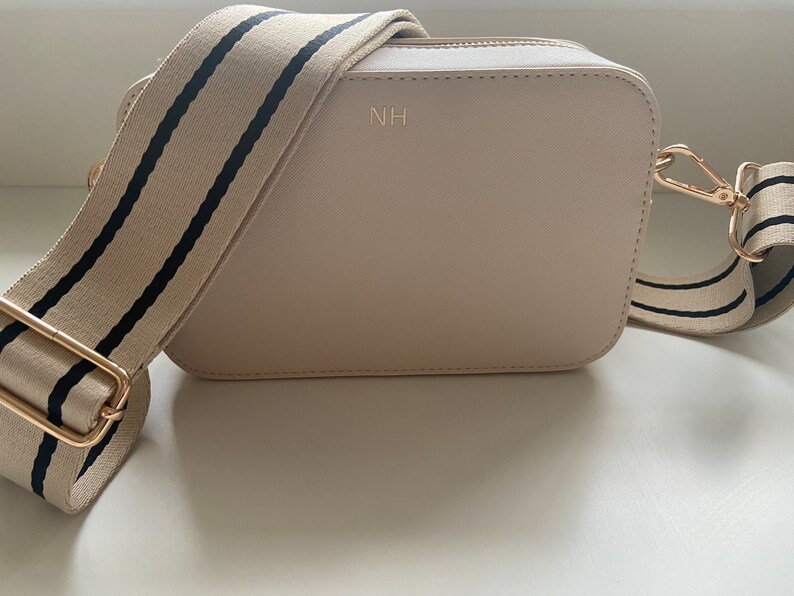 May include: A beige crossbody bag with a black and white striped strap. The bag has a gold zipper and a gold metal clasp. The initials "NH" are embossed on the front of the bag.